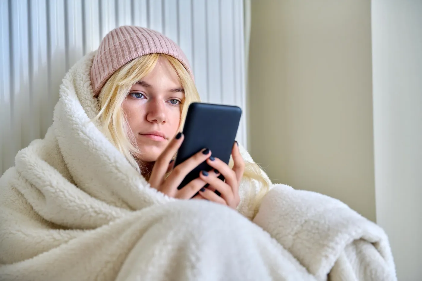 In eine Decke gehüllte Mieterin vor dem Heizkörper, Telefon in der Hand